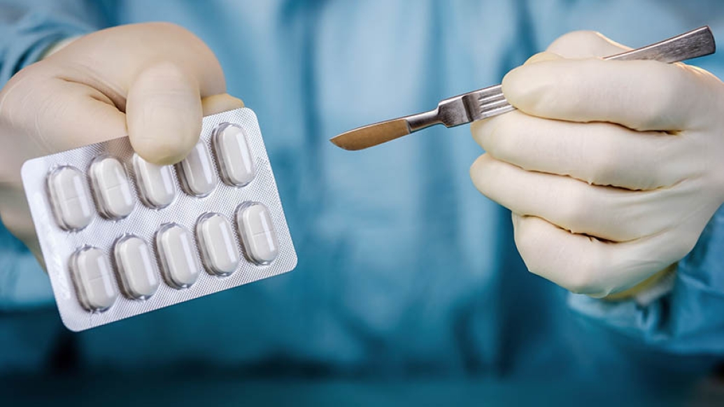 surgeon in blue scrubs holding pack of pills and scapel in other hand with white gloves on