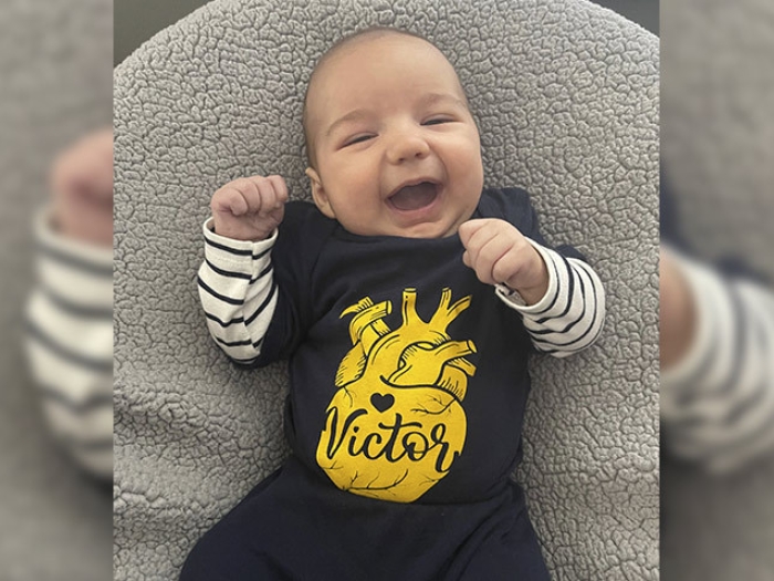 baby smiling on grey pillow with blue and yellow shirt on and striped undersleeve shirt