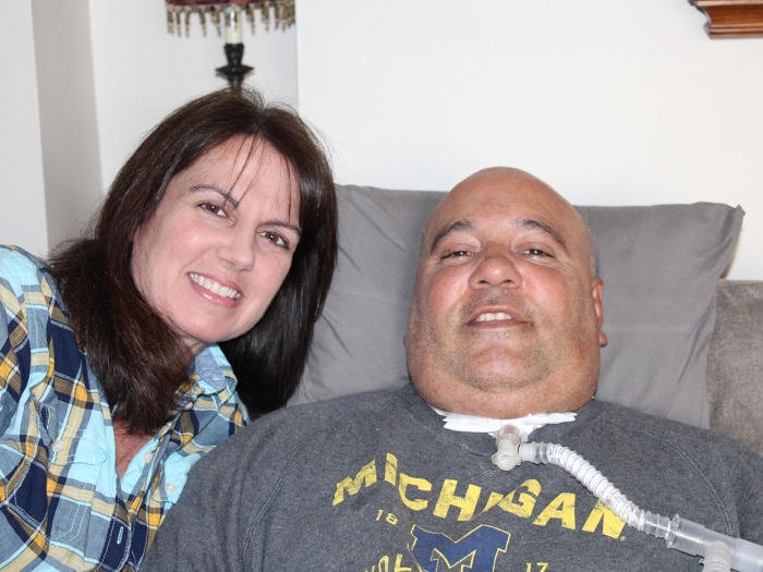 woman with dark hair standing next to man with tracheostomy sitting in chair