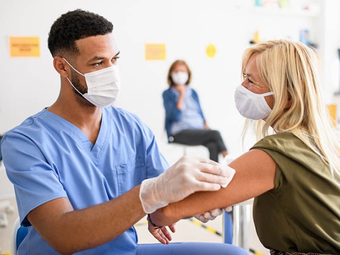 health care worker in blue scrubs and mask giving woman with blonde hair and mask a shot in her arm