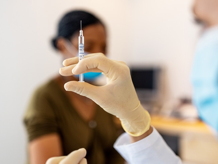 health worker holding shot in focus with mask on with patient with mask sitting in background with mask on