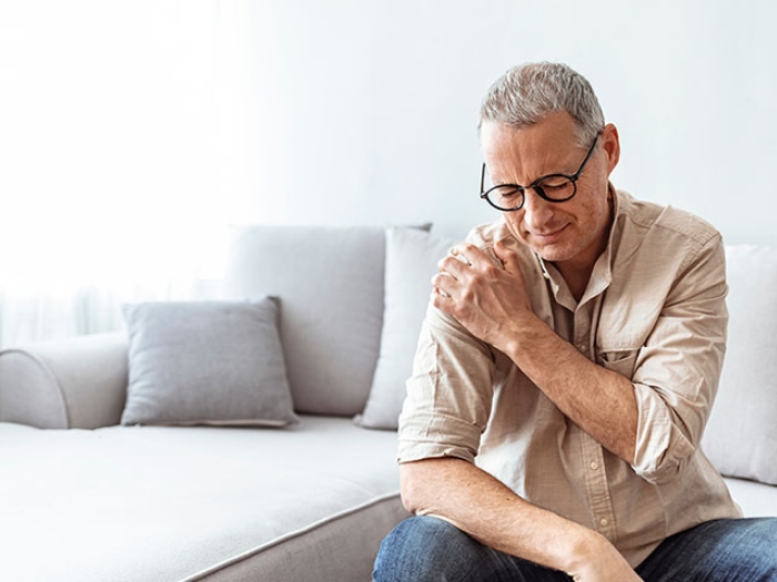 Man with glasses sitting on edge of furniture leaning, holding his shoulder.