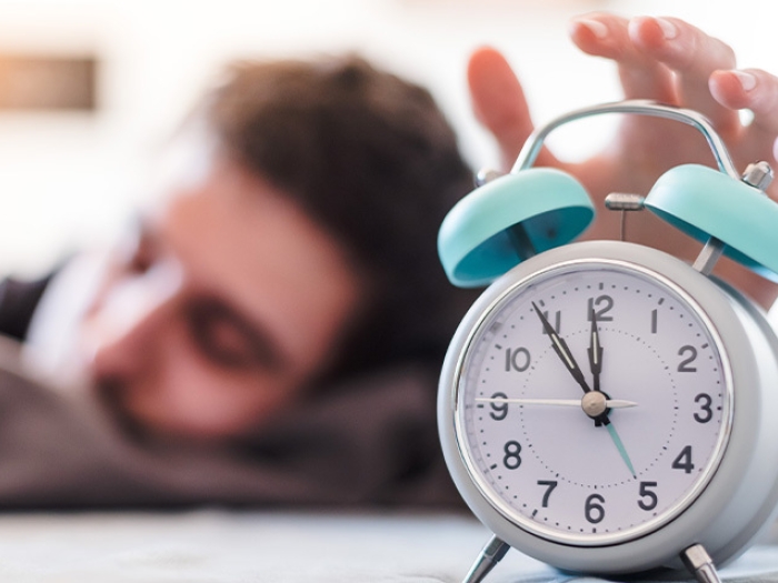 man sleeping reaching to shut off blue and white alarm clock