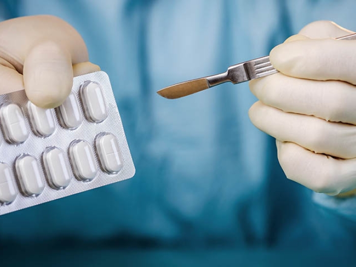 surgeon in blue scrubs holding pack of pills and scapel in other hand with white gloves on